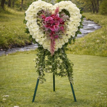 Heart-shaped standing wreath of white flowers with pink lilies and cascading green foliage on an easel outdoors.