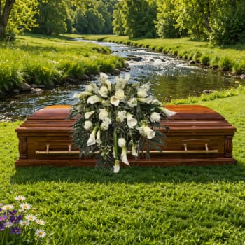 White rose and calla lily casket spray on a wooden coffin beside a grassy riverside
