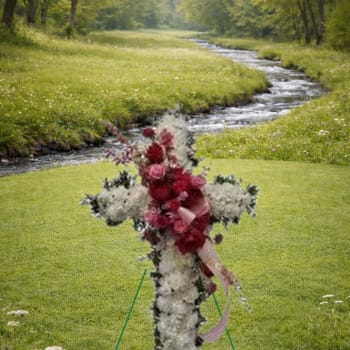 Standing floral cross of white flowers with red blooms and ribbon on an easel in a grassy outdoor setting