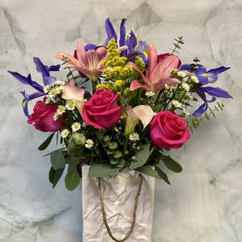Mixed bouquet of pink roses, lilies, and purple irises in a white ceramic bag-style vase