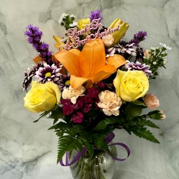 Mixed bouquet with orange lily, yellow roses, and daisies in a glass vase with a thank-you pick.