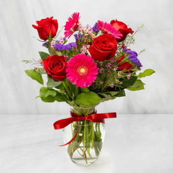 Red roses and pink gerbera daisies in a glass vase with a red ribbon