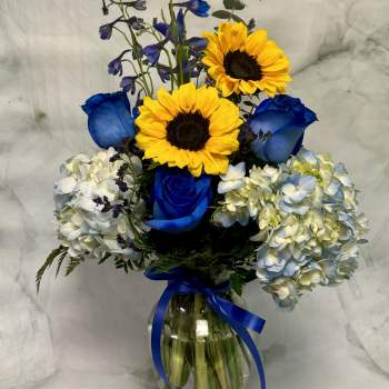 Blue roses and sunflowers in a glass vase with hydrangeas and a ribbon