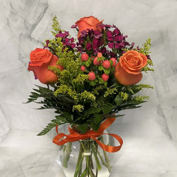 Orange roses and magenta flowers in a clear glass vase with an orange ribbon