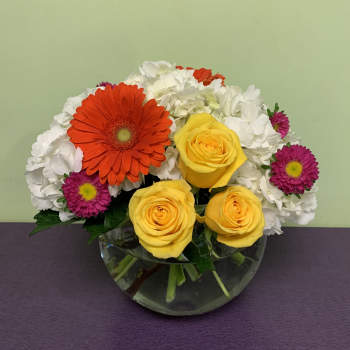 Mixed bouquet of yellow roses, orange gerbera daisies, and white blooms in a glass bowl vase