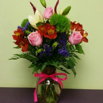 Mixed bouquet of pink roses, lilies, and red flowers in a glass vase with a pink ribbon.