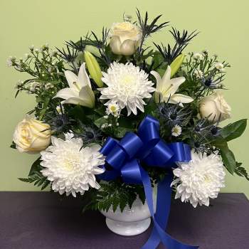 White roses and lilies arranged with chrysanthemums in a white vase with a blue ribbon