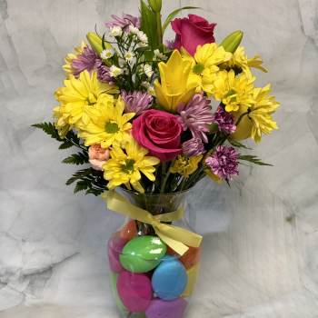 Bouquet of yellow daisies, pink roses, and lilies in a glass vase with colorful eggs