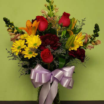 Mixed bouquet of roses, lilies, and yellow daisies in a glass vase with a lavender bow