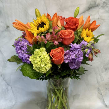 Mixed bouquet of roses, sunflowers, lilies, and purple blooms in a glass vase