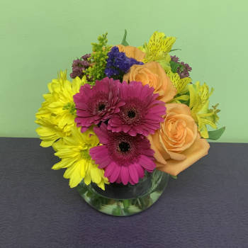 Mixed bouquet of pink gerbera daisies, yellow blooms, and peach roses in a glass vase