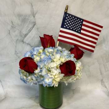 Red roses and blue-white hydrangeas in a green vase with a small U.S. flag