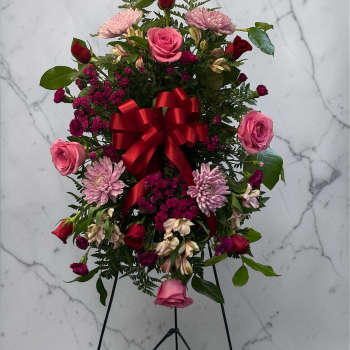 Standing spray of pink and red flowers with a red bow on a black easel.