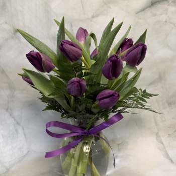 Purple tulips arranged in a clear glass vase with a purple ribbon.