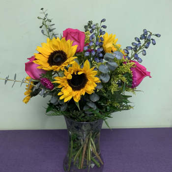 Bouquet of sunflowers, pink roses, and purple accents in a glass vase