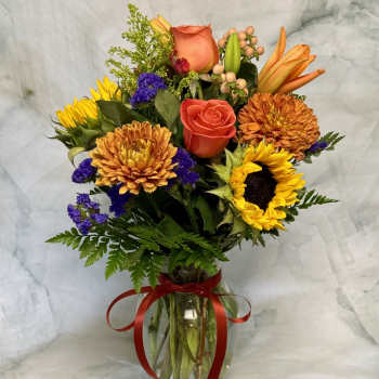 Mixed bouquet of sunflowers, roses, and orange blooms in a glass vase with a red ribbon