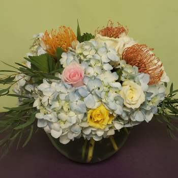 Mixed bouquet of hydrangeas, roses, and orange pincushion blooms in a glass vase
