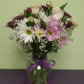 Mixed bouquet of daisies, roses, and purple blooms in a glass vase with ribbon