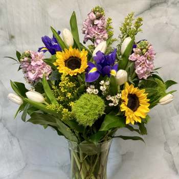Mixed bouquet with sunflowers, purple irises, white tulips, and pink blooms in a glass vase