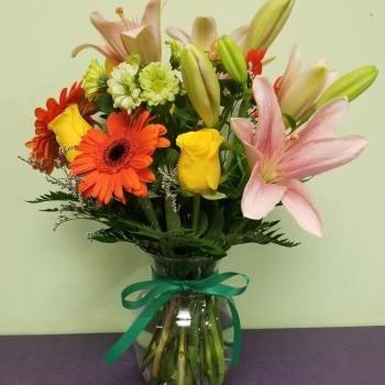 Mixed bouquet of lilies, roses, and gerbera daisies in a glass vase with ribbon