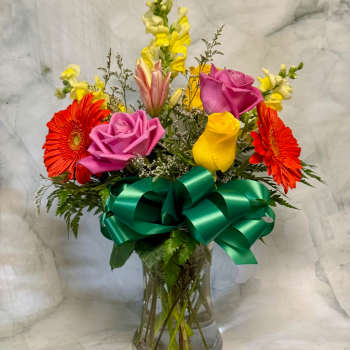 Mixed bouquet of pink, yellow, and orange flowers in a clear vase with a large green ribbon bow