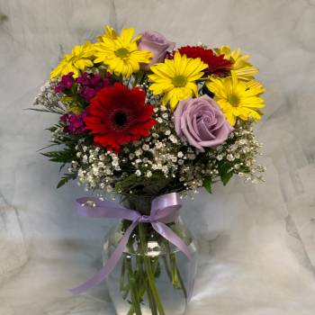 Mixed bouquet of yellow daisies, red gerbera daisies and lavender roses in a clear vase with a purple ribbon