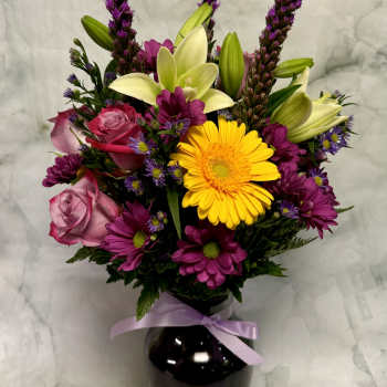 Mixed bouquet of roses, lilies, and daisies in a black vase with a lavender ribbon