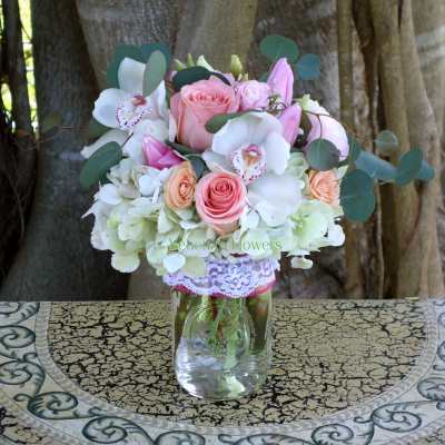 Mason jar arrangement with pink and peach roses, white orchids, hydrangeas, and pink lilies.