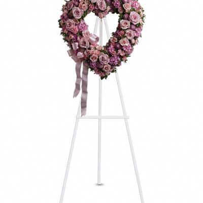 Heart-shaped wreath of pink and lavender roses on a white standing easel with a satin bow.