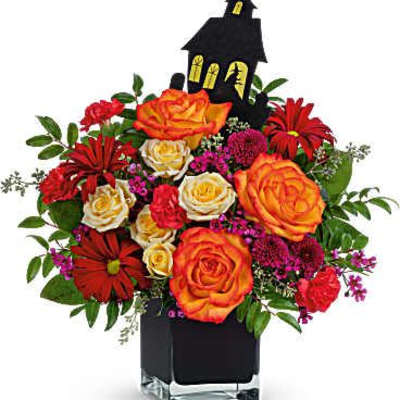 Orange and red roses with red daisies in a black vase topped by a haunted house decoration