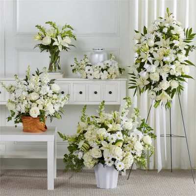 Collection of white funeral flower arrangements with lilies and roses around an urn and standing spray.