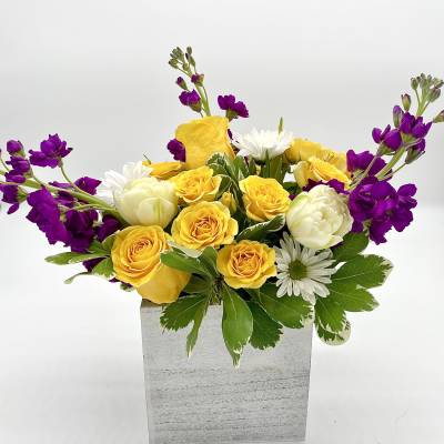 Yellow roses and white daisies in a square gray vase with purple flowers