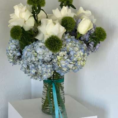 White roses and blue hydrangeas in a glass vase with green pom-pom accents