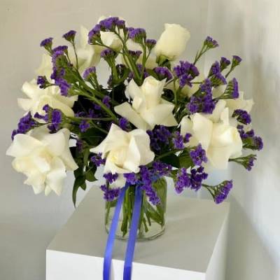 White roses and purple filler flowers in a clear glass vase with blue ribbon