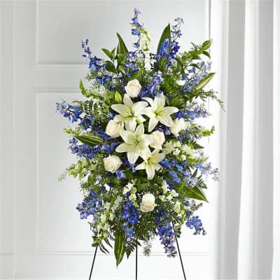Standing spray of white lilies and roses with blue delphinium on a metal easel.