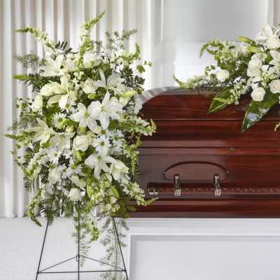Tall white funeral spray with lilies, roses, and daisies on a stand beside a wooden casket.