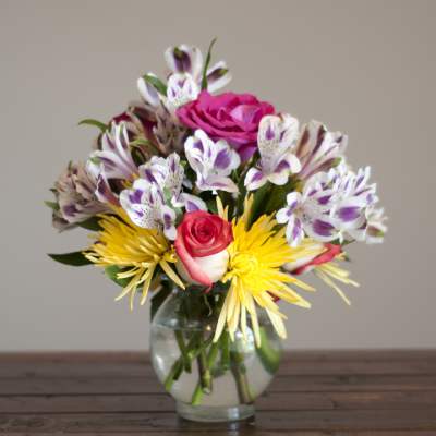 Mixed bouquet of pink roses, yellow spider mums, and white-purple alstroemeria in a glass vase