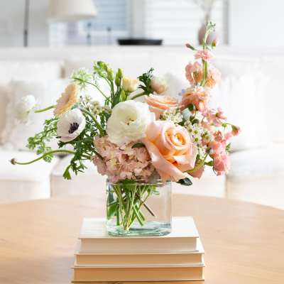 Pastel arrangement of peach roses and white blooms in a glass cube vase on books on a coffee table