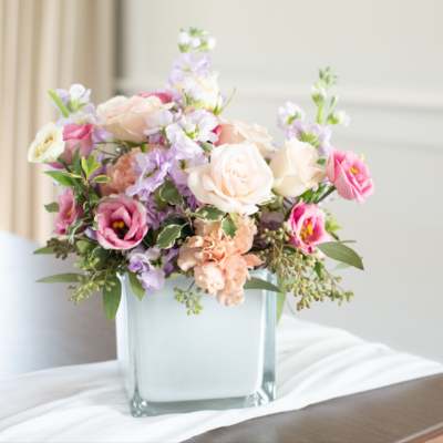 Low pastel arrangement of roses and lisianthus in a frosted glass cube vase on a draped table