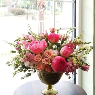 Low gold compote of bright pink peonies, peach roses, and white blooms on a table by a window