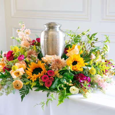 Cremation urn encircled by bright sunflowers, roses, and mixed blooms in a low table arrangement