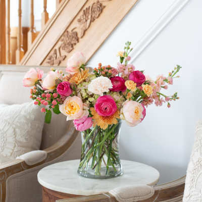 Mixed bouquet of pink, peach, and yellow flowers in a clear vase on a small table between chairs