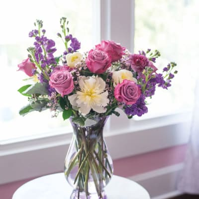 Mixed bouquet of pink roses, white peonies, and purple flowers in a clear lavender glass vase on a small table
