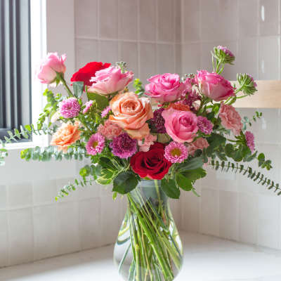 Mixed bouquet of pink, peach, and red roses with small mums in a clear glass vase