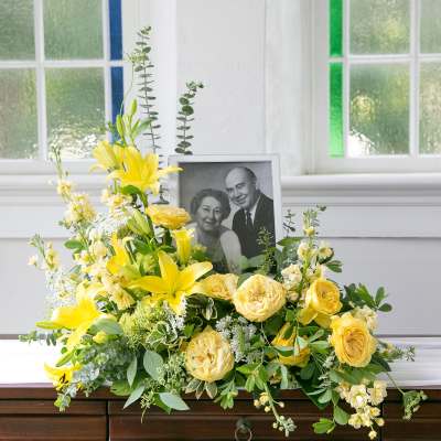 Yellow lily and rose sympathy arrangement displayed with a framed black-and-white portrait.