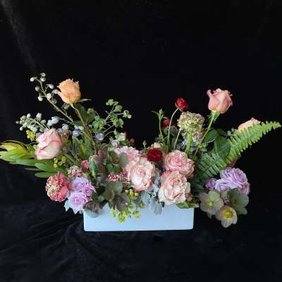 Low pastel arrangement of peach roses, lavender flowers, and green accents in a white rectangular vase