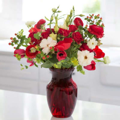 Red and white mixed flower arrangement with berries in a red glass vase on a white countertop