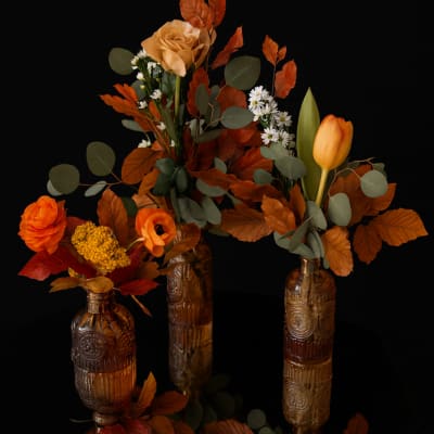 Three autumn floral arrangements in amber glass bottles with orange blooms