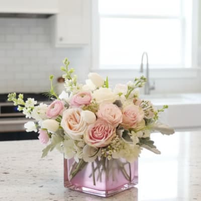 Low pastel pink and white flower arrangement in a pink glass cube vase on a kitchen counter