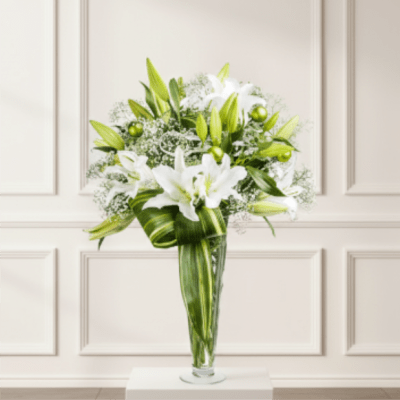 Tall glass vase of white lilies with baby's breath and broad green leaves in a modern arrangement.